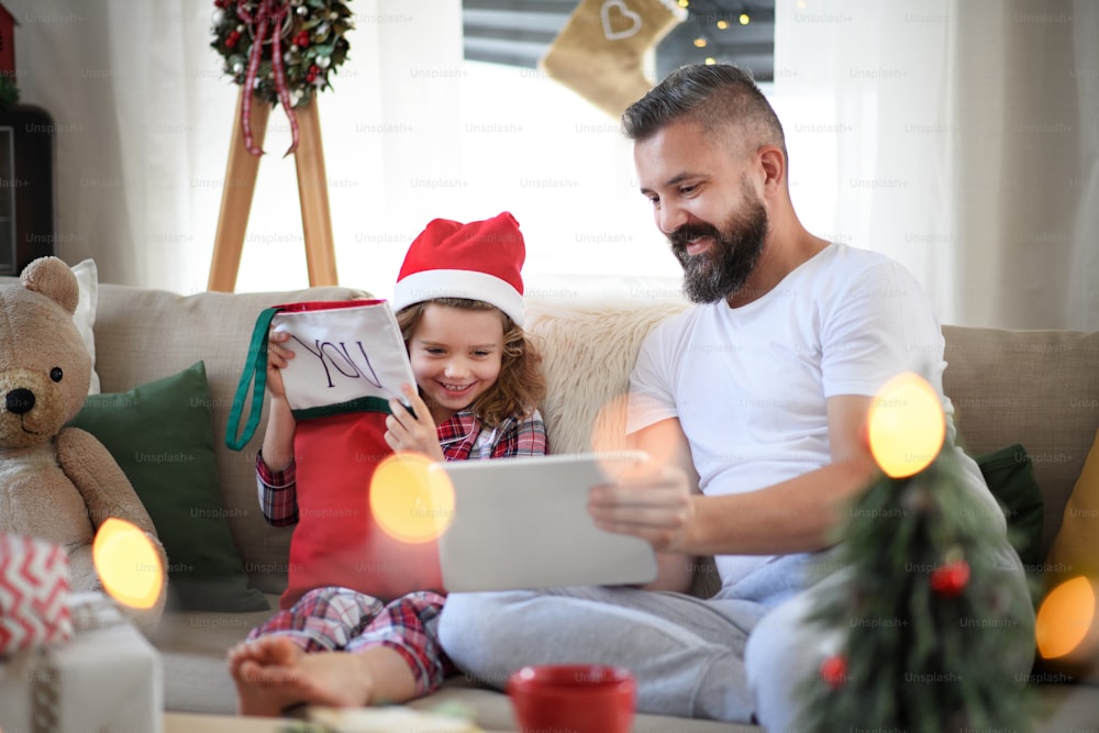 Portrait d’un père avec sa petite fille à l’intérieur à la maison à Noël, à l’aide d’une tablette.