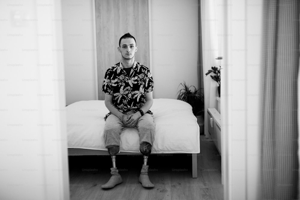 A black and white portrait of disabled young man sitting on bed indoors at home, leg prosthetic concept.