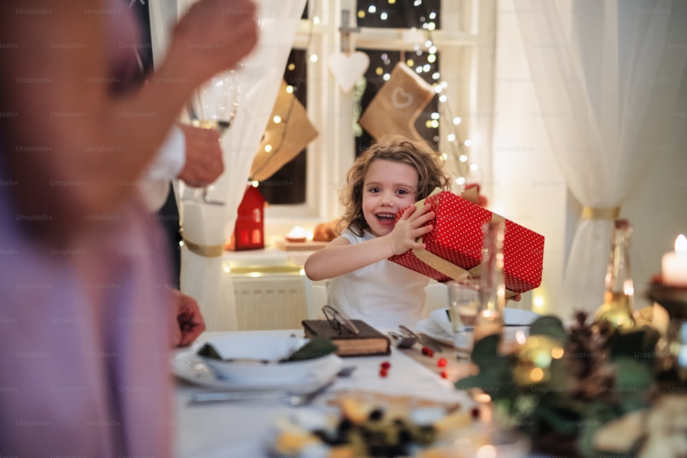 Petite fille heureuse avec sa famille à la table à l’intérieur célébrant Noël ensemble, tenant un cadeau.