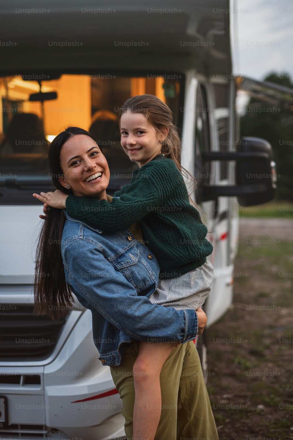 Madre feliz con hija de pie junto al coche al aire libre en el camping al anochecer, viaje de vacaciones familiares en caravana.