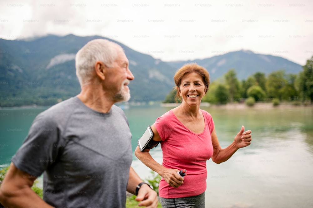 A senior pensioner couple with smartphone running by lake in nature.