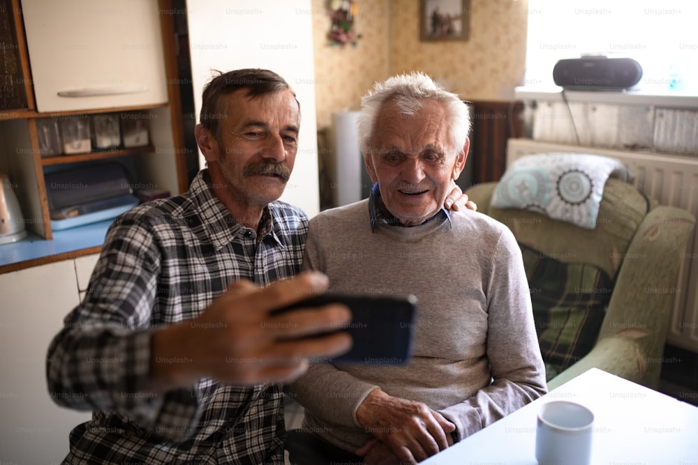 A portrait of man with elderly father sitting at the table indoors at home, taking selfie.