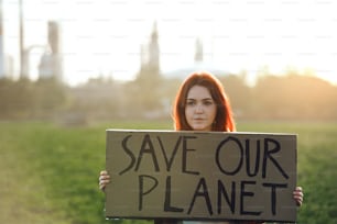 Portrait of young woman activist with placard standing outdoors by oil refinery, protesting.