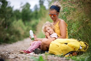 Mother with small toddler daughter hiking outdoors in summer nature, resting and drinking water.
