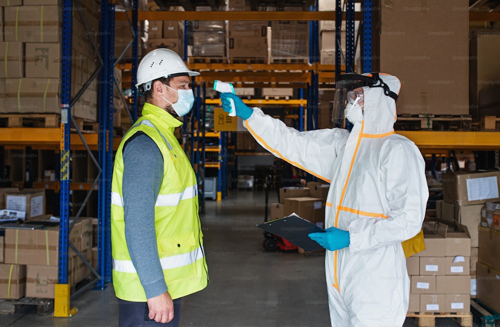 Lavoratori con maschera facciale e tuta protettiva in magazzino, coronavirus e concetto di misurazione della temperatura.