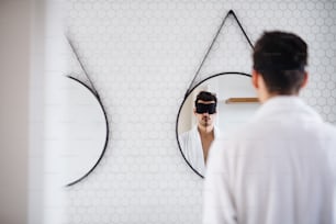 Jeune homme avec un masque pour les yeux dans la salle de bain le matin, debout devant un miroir.