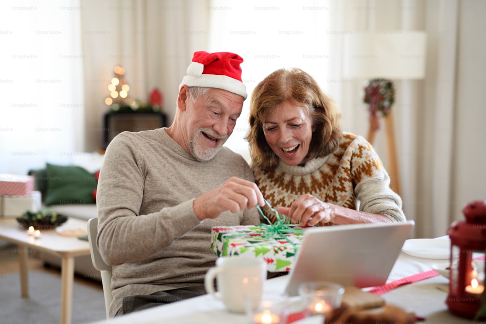 Feliz casal de idosos abrindo presente dentro de casa no Natal, tendo chamada de vídeo com a família.