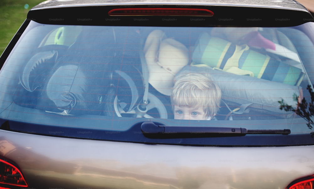 Small boy sitting in loaded car boot with closed door, local trip concept. Shot through glass.
