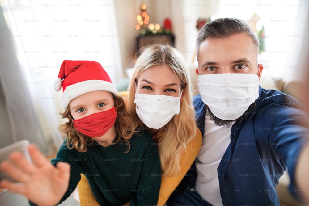 Vue de face d’une famille avec une petite fille à l’intérieur à la maison à Noël, prenant un selfie avec des masques faciaux.