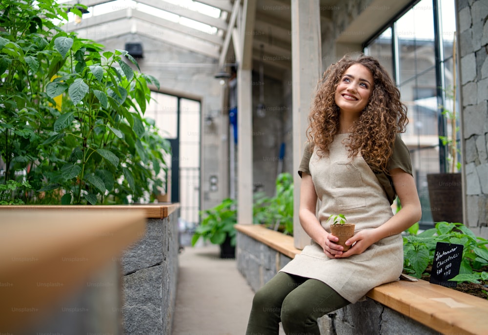Portrait of woman gardener sitting in greenhouse, holding small plant.