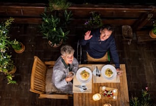 Draufsicht eines älteren Paares im Rollstuhl, das abends auf der Terrasse zu Abend isst und in die Kamera schaut.
