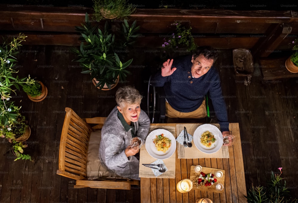 Vue de dessus d’un couple de personnes âgées en fauteuil roulant dînant le soir sur la terrasse, regardant la caméra.