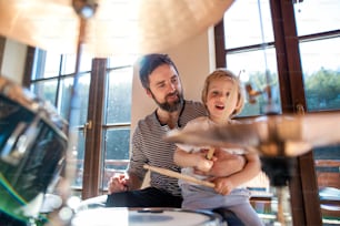 Front view of portrait of small boy with father indoors at home, playing drums.