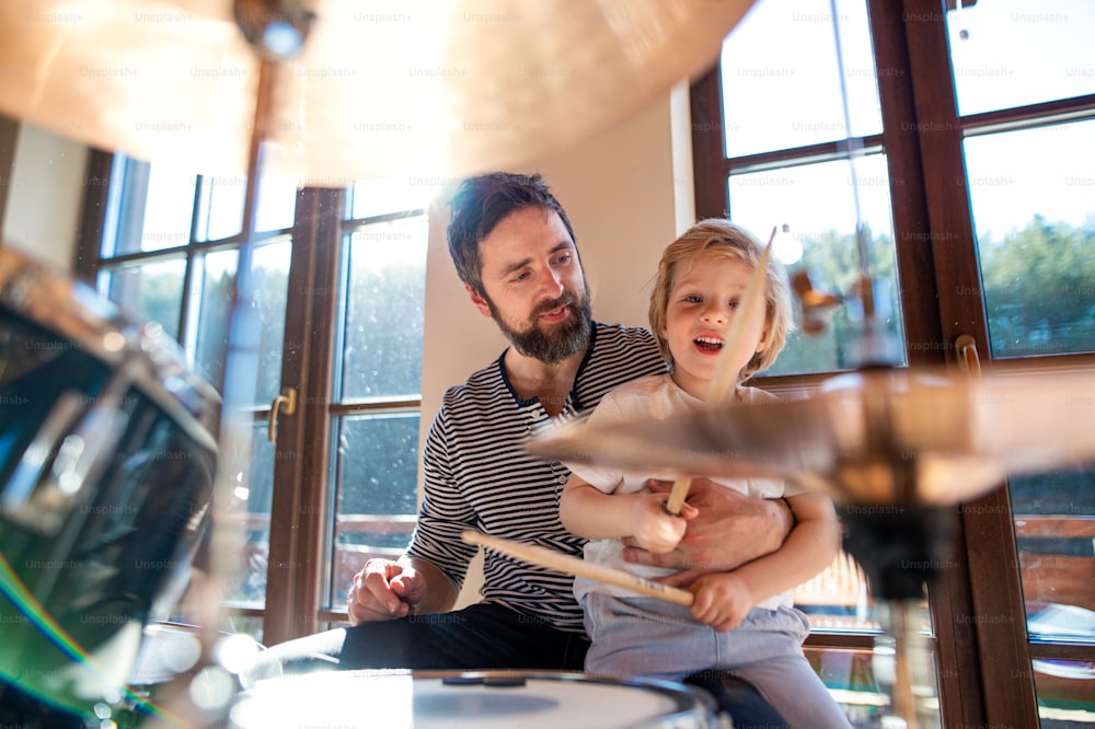 Vista frontal del retrato de un niño pequeño con el padre en el interior de la casa, tocando la batería.