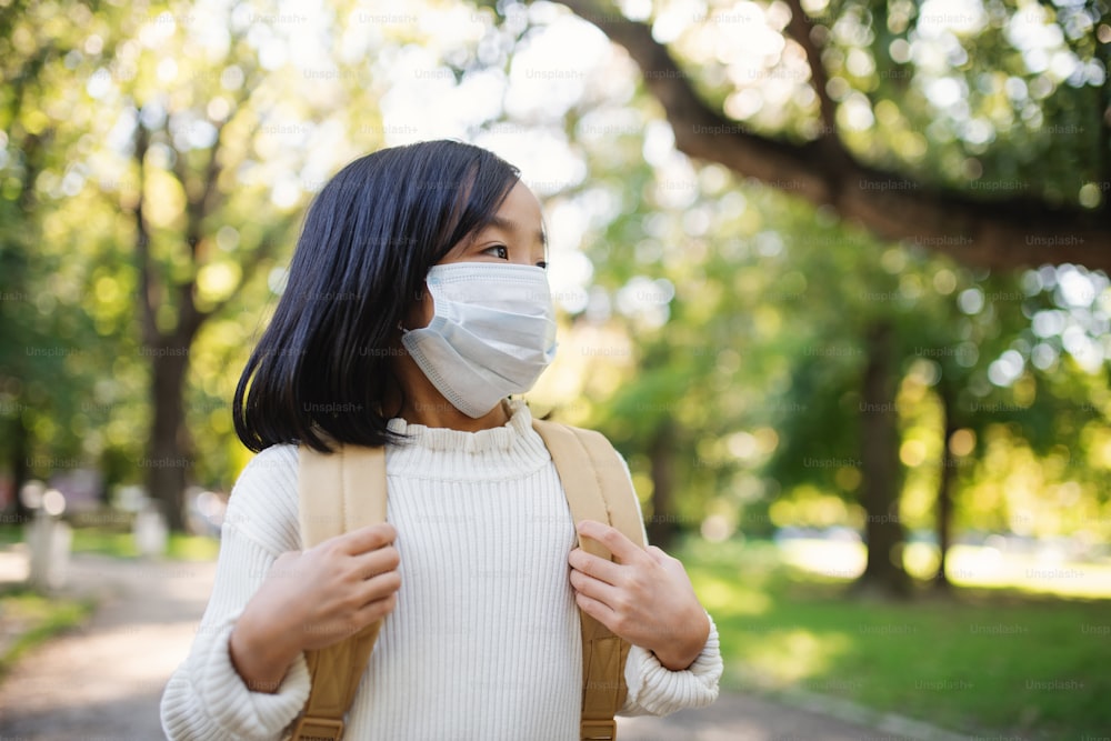 Portrait de vue de face d’une petite fille japonaise avec un sac à dos marchant à l’extérieur en ville, concept de coronavirus.