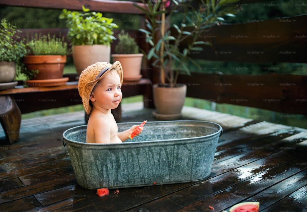 Ragazzino felice con un cappello nella vasca da bagno all'aperto in giardino in estate, mangiando anguria.