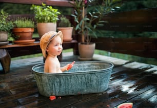 Happy small boy with a hat in bath tub outdoors in garden in summer, eating watermelon.