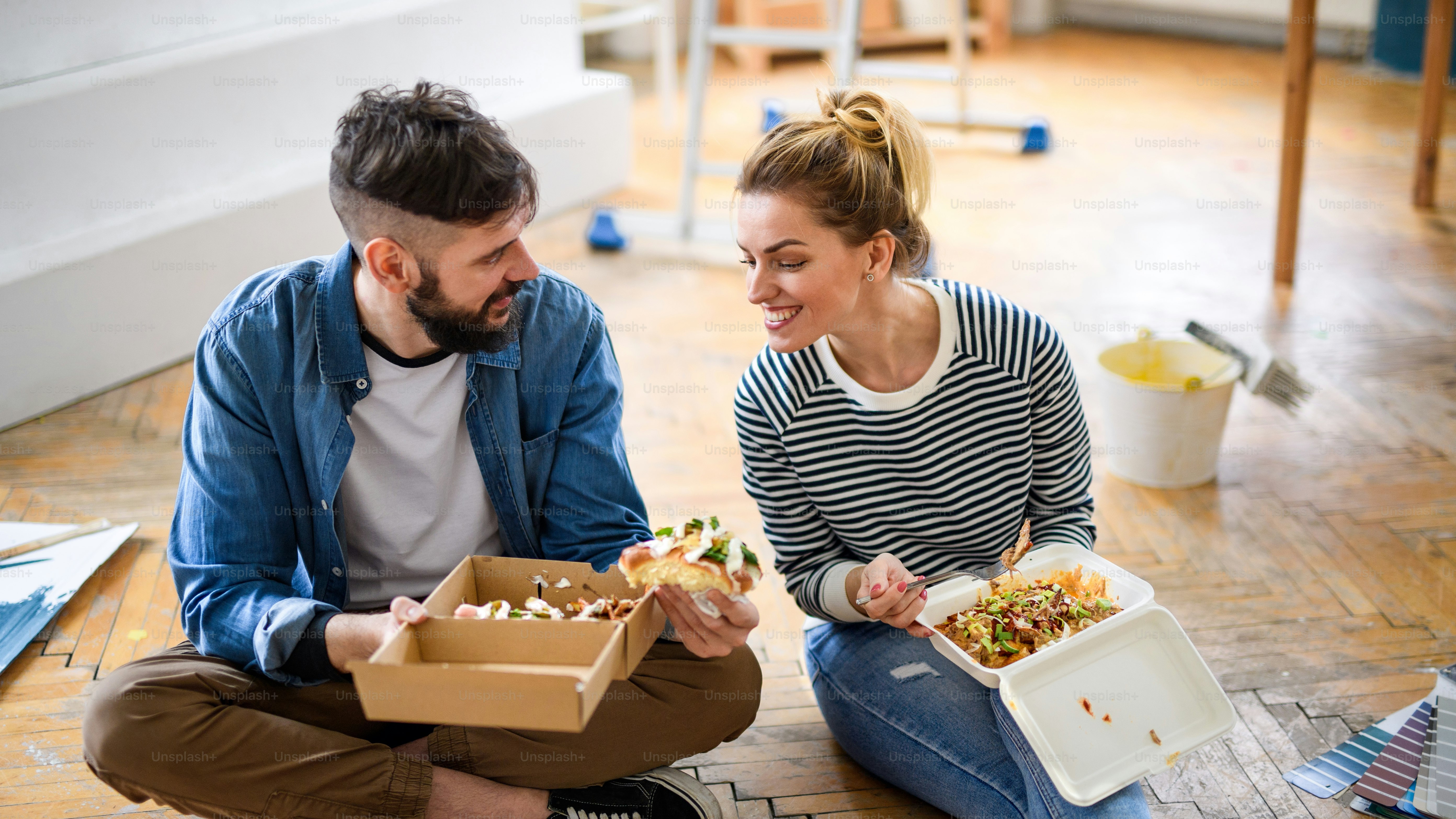 Mid adults couple eating lunch indoors at home, relocation, diy and food delivery concept.