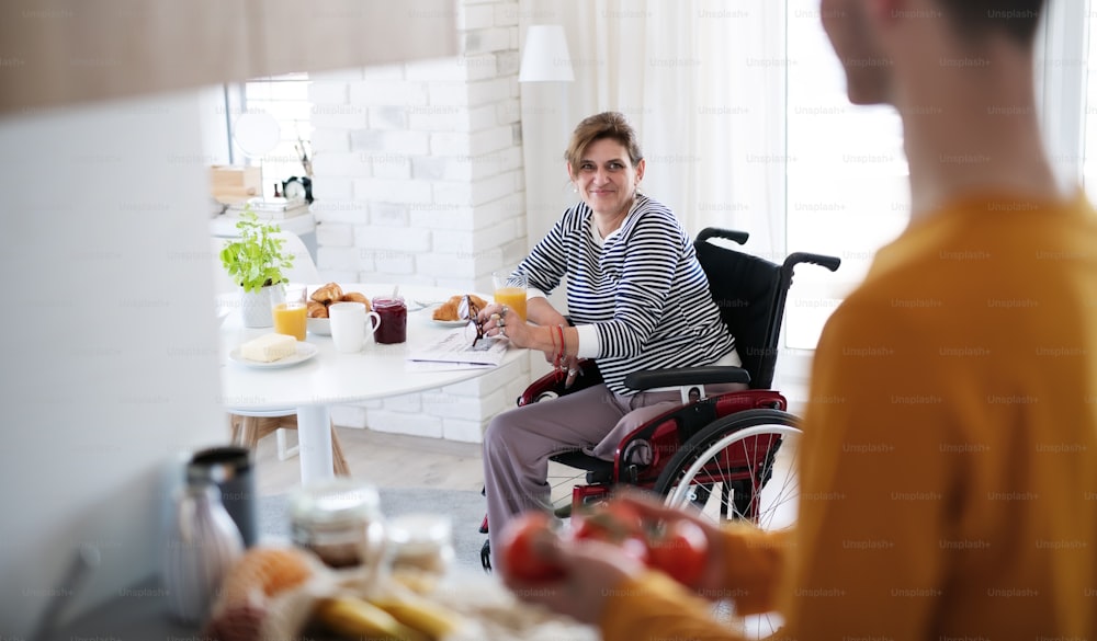 Una donna matura disabile in sedia a rotelle seduta al tavolo all'interno di casa, parlando con un amico.
