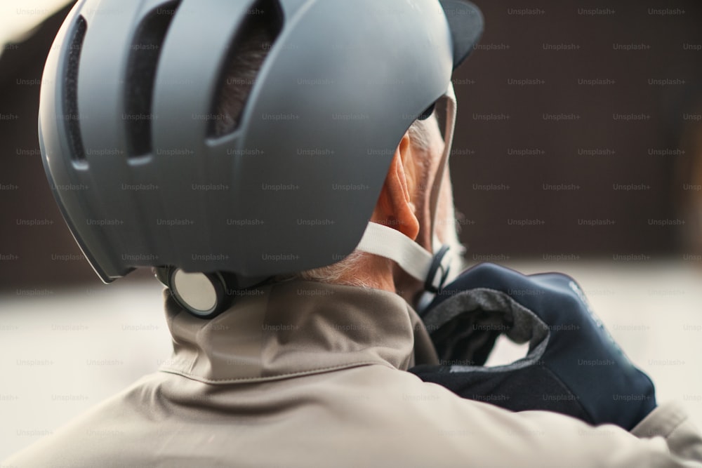 Uma visão traseira de um homem idoso ativo em pé ao ar livre na cidade, colocando um capacete de bicicleta. Um close-up.