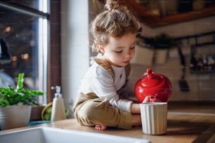 Cute small toddler girl sitting on kitchen counter indoors at home, pouring tea in cup.