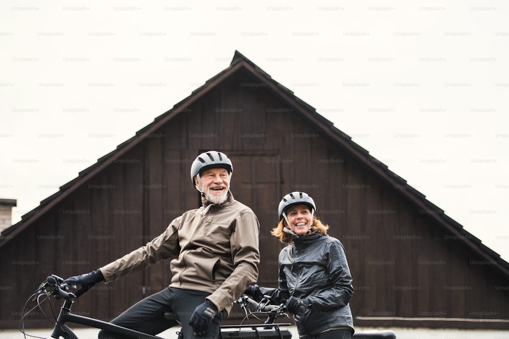 Un couple de personnes âgées actives avec des casques et des vélos électriques debout à l’extérieur devant une maison.