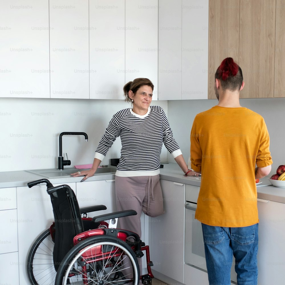 A disabled mature woman in wheelchair talking to son in kitchen indoors at home.