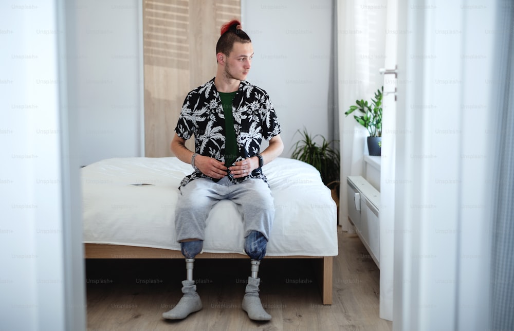 Retrato da vista frontal do jovem deficiente sentado na cama dentro de casa, conceito protético de perna.
