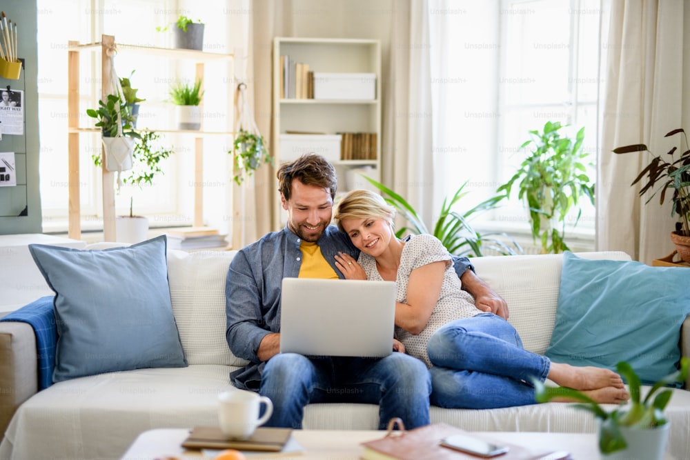 Vista frontal de una pareja feliz enamorada sentada en el interior de la casa, usando una computadora portátil.