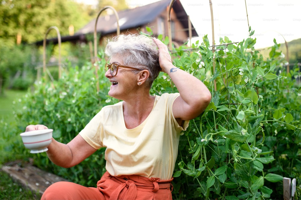 Portrait of contented senior woman sitting outdoors by vegetable garden, holding cup of coffee.