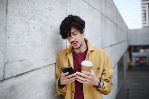A portrait of young man standing against concrete wall outdoors, using smartphone.