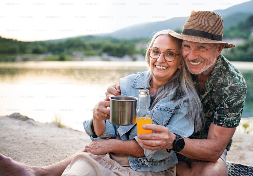 A happy senior couple on hiking trip on summer holiday, relaxing by lake.