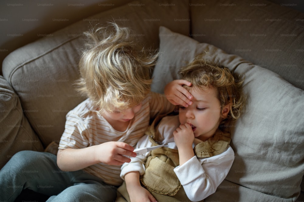 Two small sick children brother and sister at home lying in bed, measuring temperature.