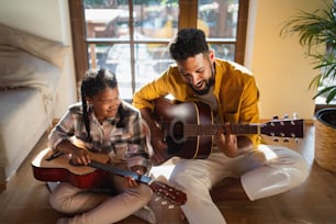 A high-angle view of happy father with small daughter indoors at home, playing guitar.
