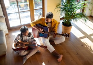 A high-angle view of happy father with small daughter indoors at home, playing guitar.