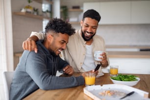 A portrait of young adult brothers in kitchen indoors at home, using smartphone when eating pizza.