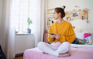 Sad young female student sitting on bed, using smartphone. Copy space.