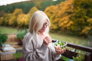 Senior woman standing outdoors on terrace, eating healthy salad snack.