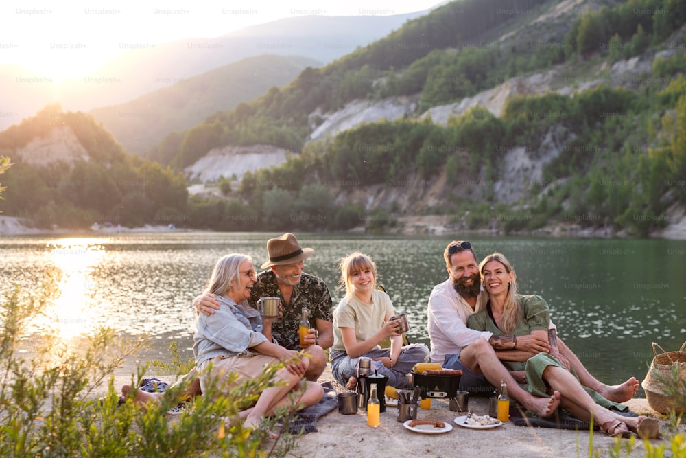 A happy multigeneration family on summer holiday trip, barbecue by lake at sunset.