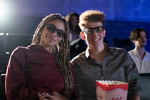 Jovem casal com óculos 3D e pipoca assistindo filme no cinema, olhando para a câmera.