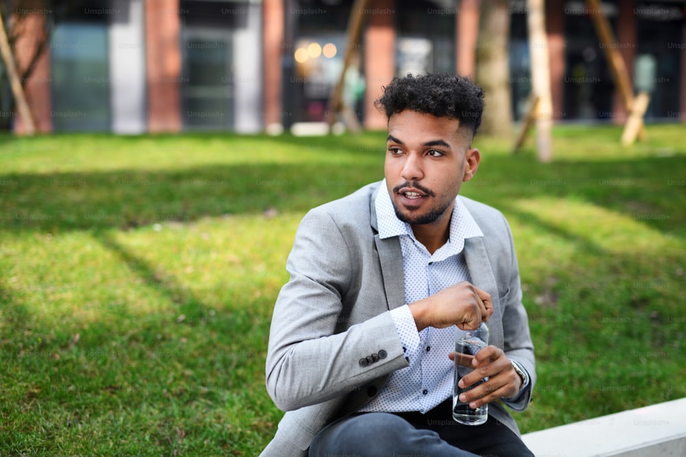 Un portrait d’un jeune homme étudiant assis à l’extérieur dans la ville, tenant une bouteille d’eau.