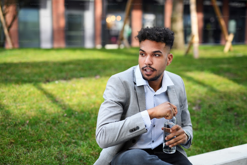 A portrait of young man student sitting outdoors in city, holding water bottle.