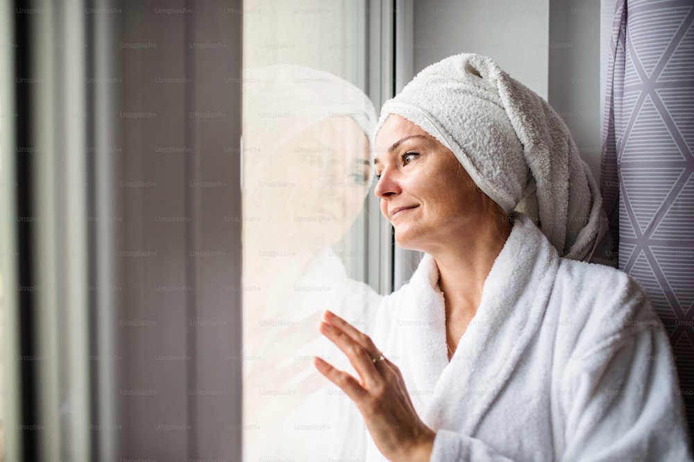 Porträt einer einsamen Frau mit Bademantel und Handtuch auf dem Kopf, die zu Hause drinnen steht.