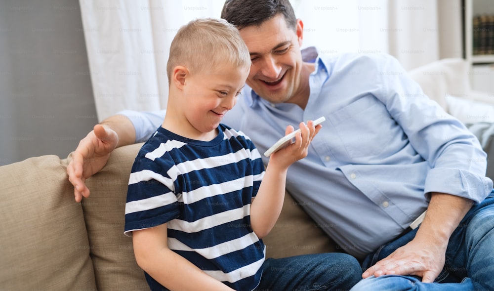 Un padre con un hijo feliz con síndrome de Down en el interior de su casa, usando un teléfono inteligente para mirar.
