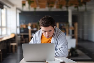 A concentrated young man with Down syndrome sitting and studying indoors at school, using laptop
