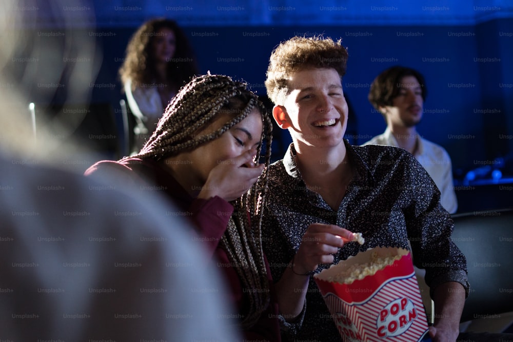 Cheerful young couple eating popcorn while watching movie in cinema, laughing.