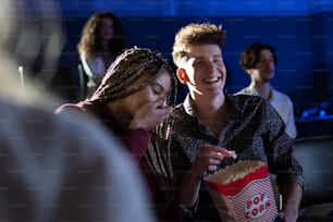 Cheerful young couple eating popcorn while watching movie in cinema, laughing.