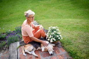 Side view portrait of senior woman with pet dog sitting outdoors in garden, relaxing with coffee.