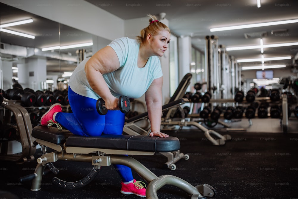 A mid adult overweight woman exercising with dumbbells on bench indoors in gym