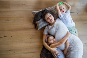 Vista superior de una mujer embarazada feliz con niños pequeños en el interior de la casa, acostada en el suelo.